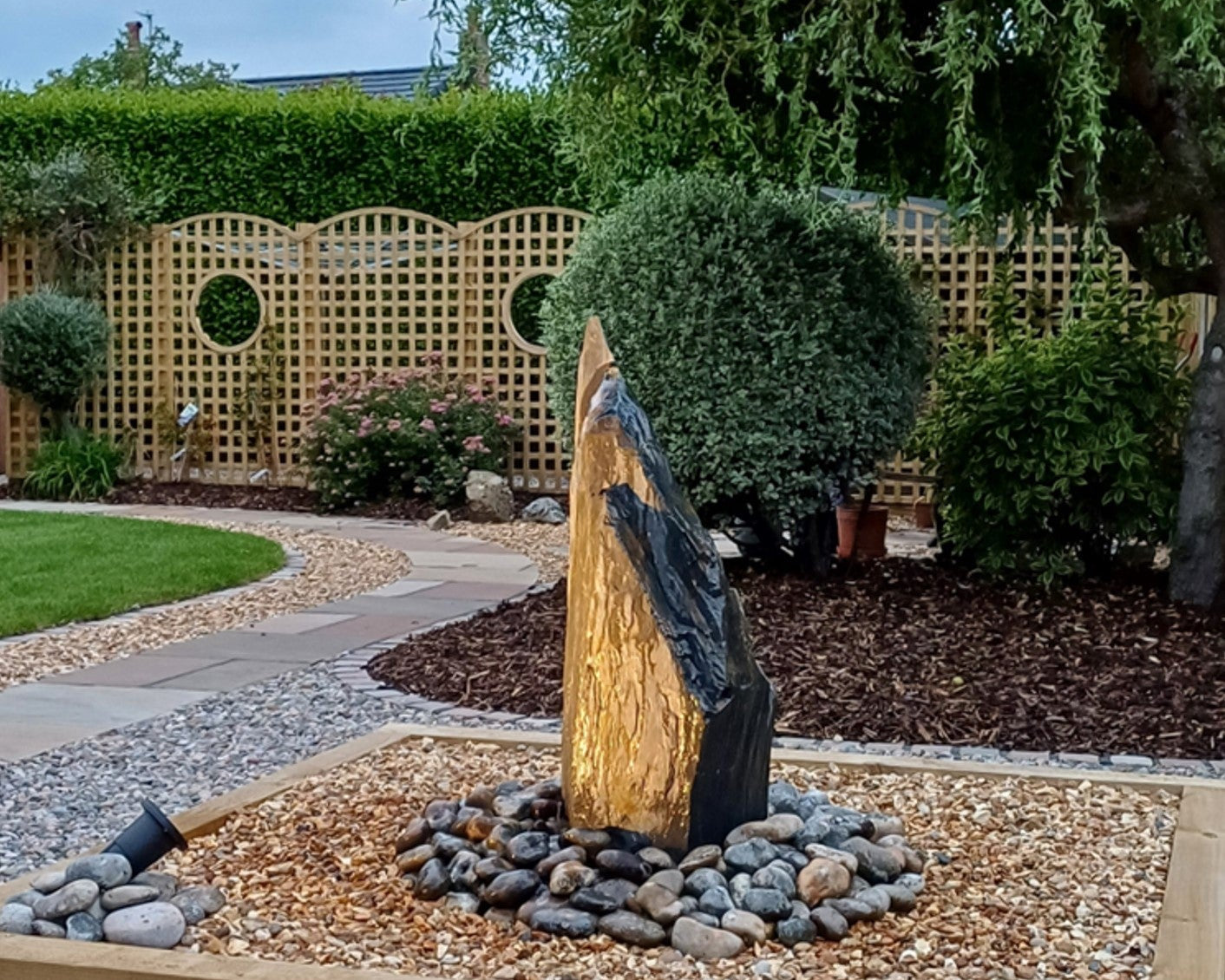 A garden scene features a stone water fountain surrounded by gravel and rocks, with a wooden lattice fence in the background.
