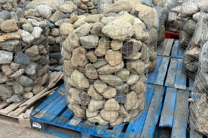 Several wire cages filled with smooth river rocks sit on blue wooden pallets in an outdoor setting.
