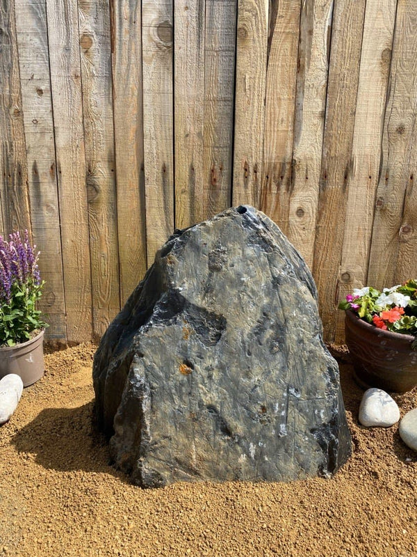 Slate monolith water feature by C09 standing on gravel with plants and a wooden fence in the background.