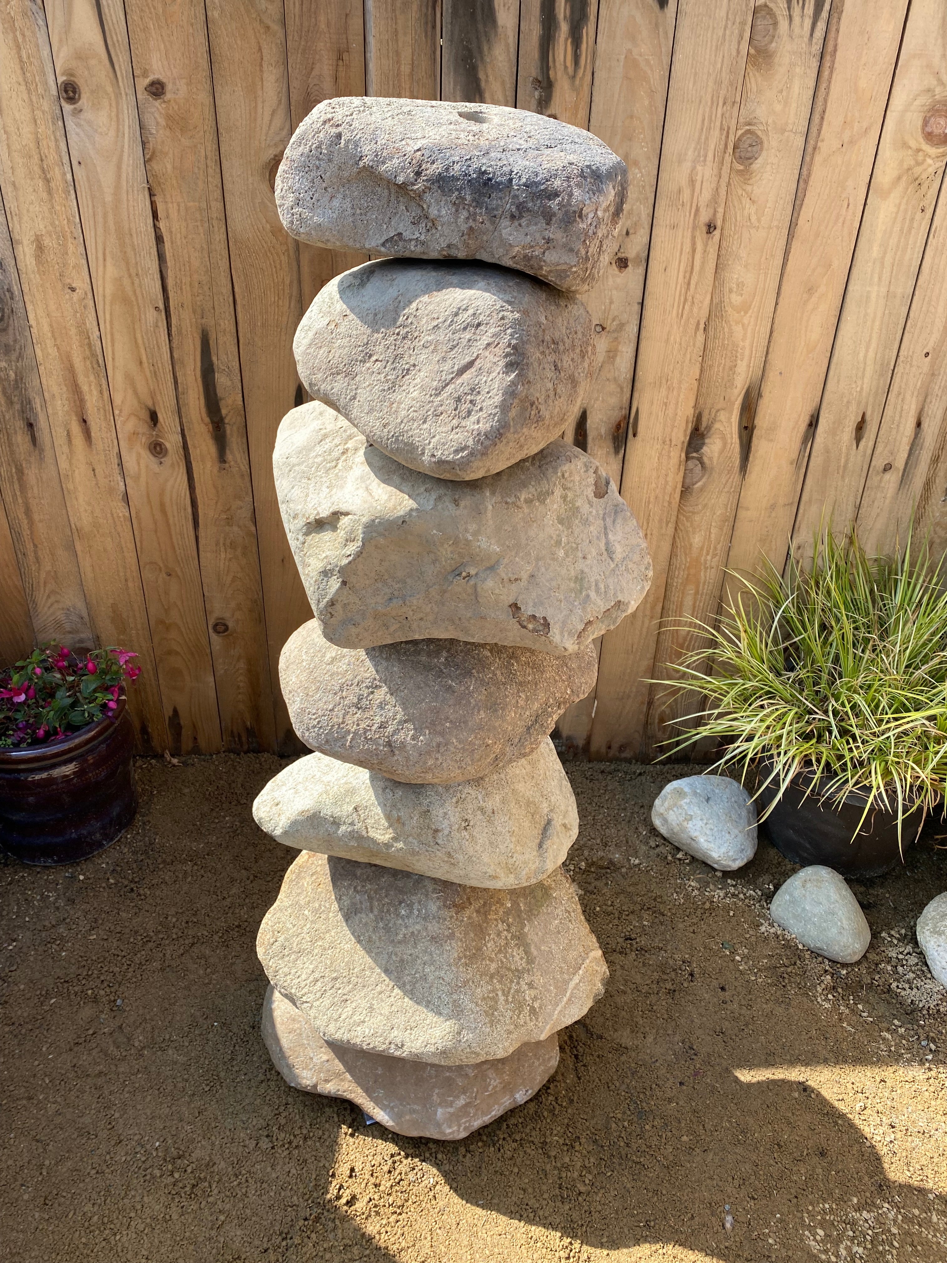 Large natural pebble water feature, one meter tall, with stacked stones against a wooden fence.