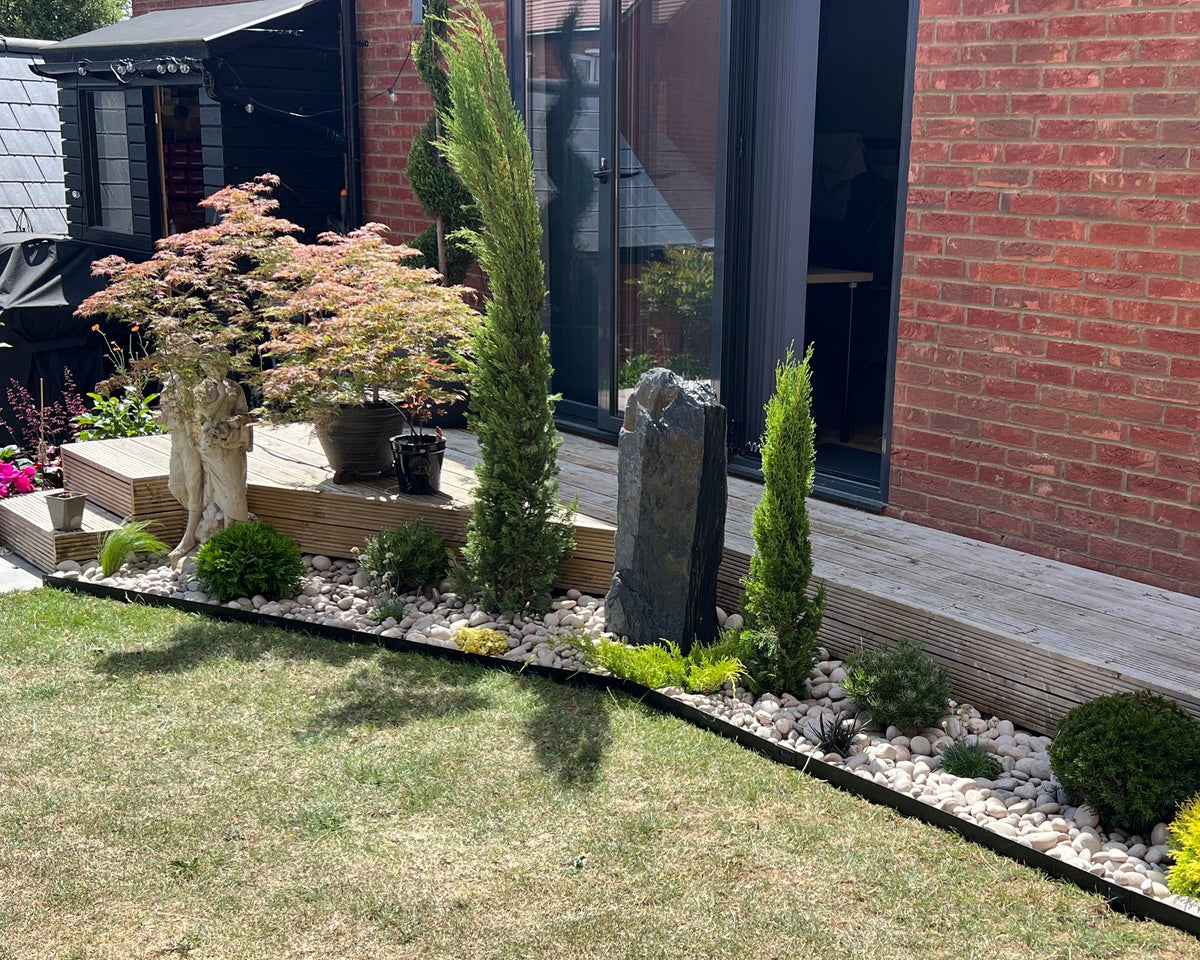 A well-maintained garden with green lawn, flowerbeds with white rocks, and a small statue on wooden decking. Brick building in background.