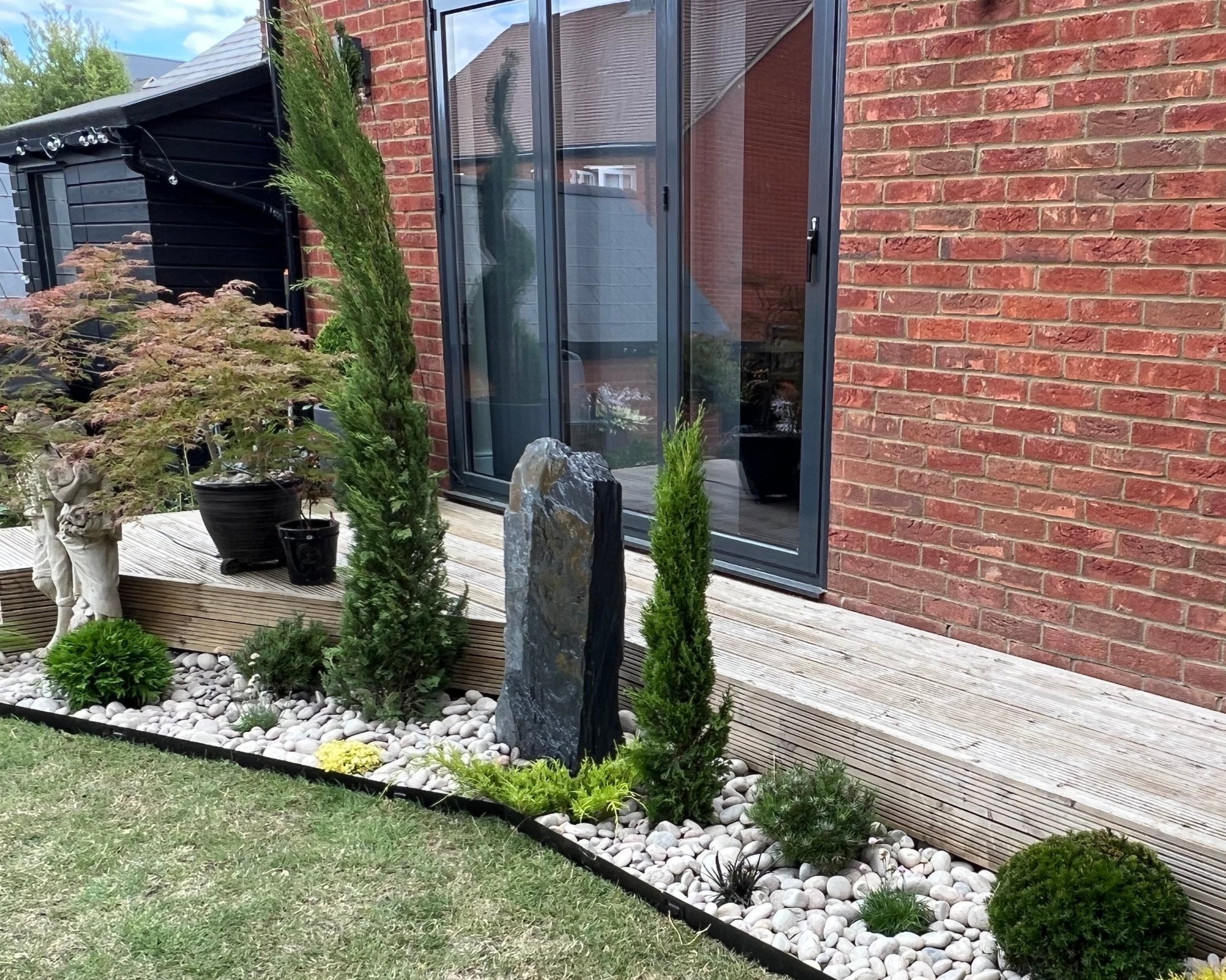 A landscaped garden bed features white pebbles, green plants, and a slate monolith alongside a brick wall and glass doors. A wooden deck adds dimension.
