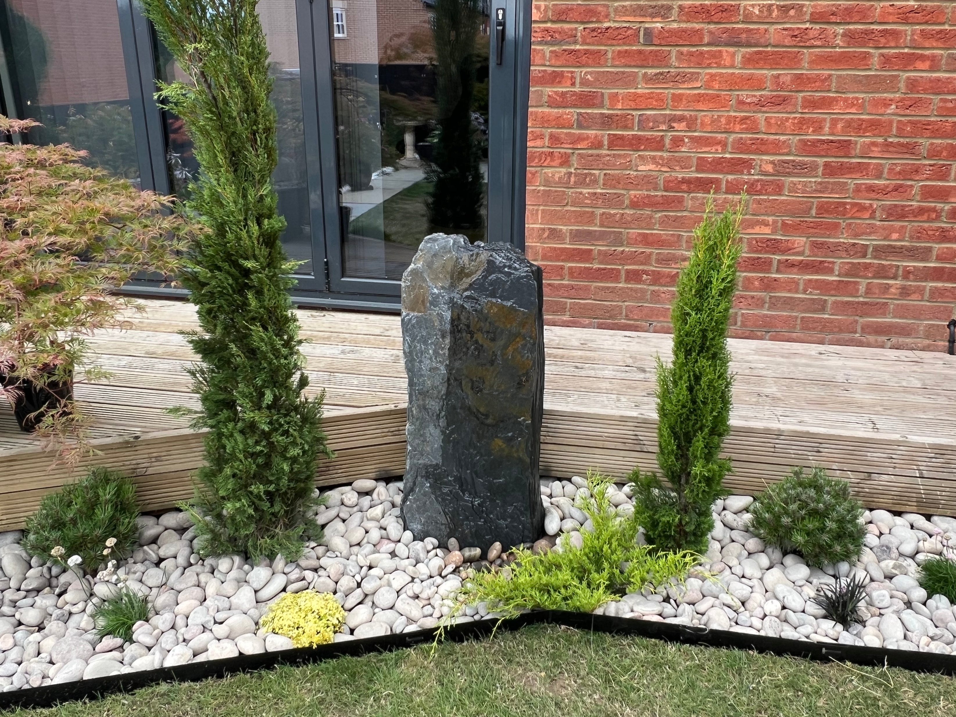 A landscaped garden bed featuring a tall, dark stone pillar surrounded by pebbles, shrubs, and two slender conifer trees against a red brick wall.