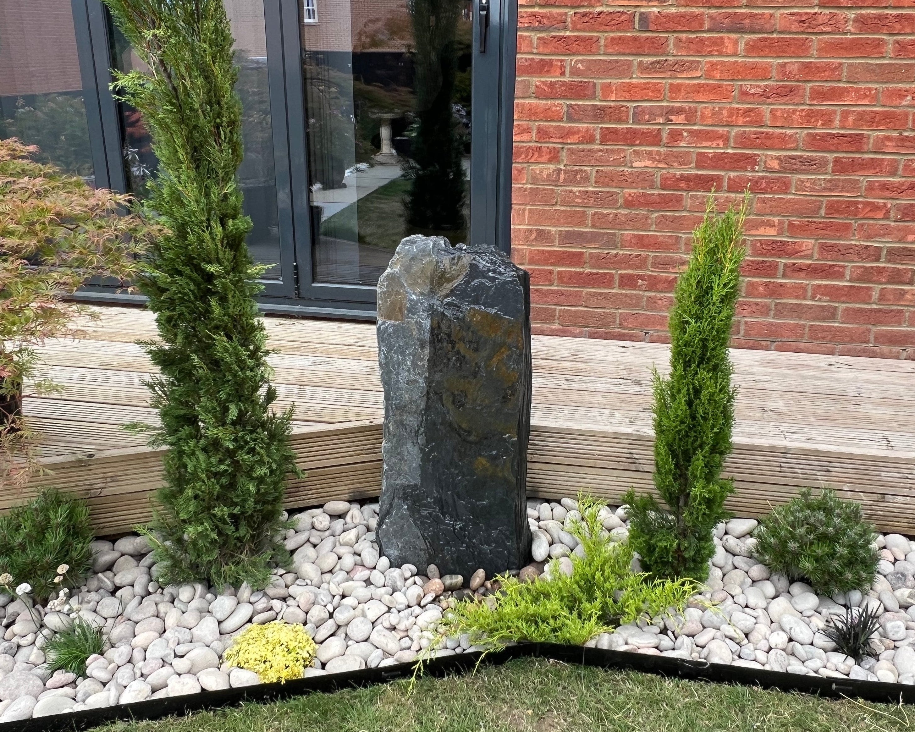 Outdoor garden scene with a dark stone centerpiece, flanked by green plants and white rocks, against a brick wall backdrop.