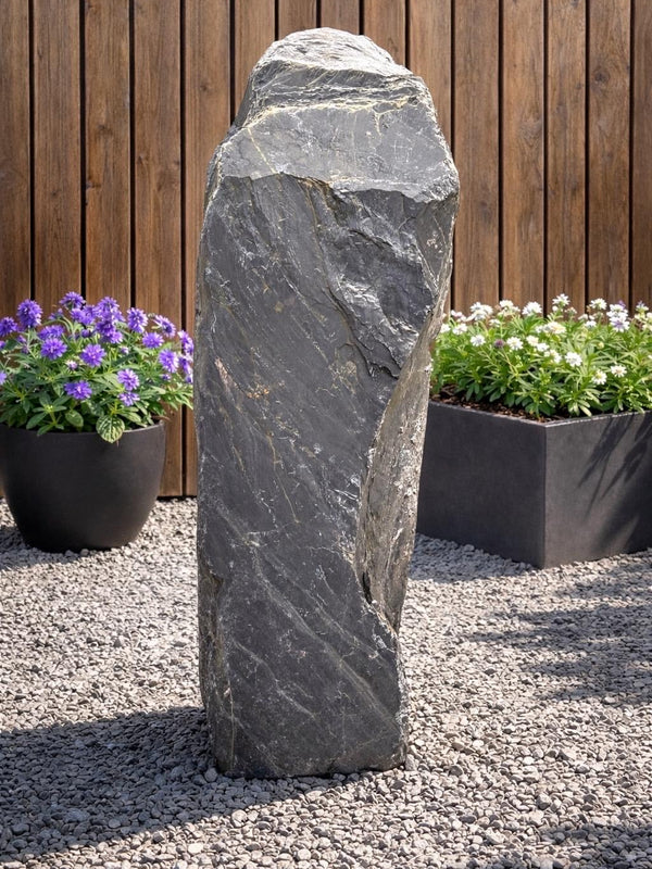 Slate monolith water feature by Water Feature 101, standing among gravel, with purple and white flowers in the background.