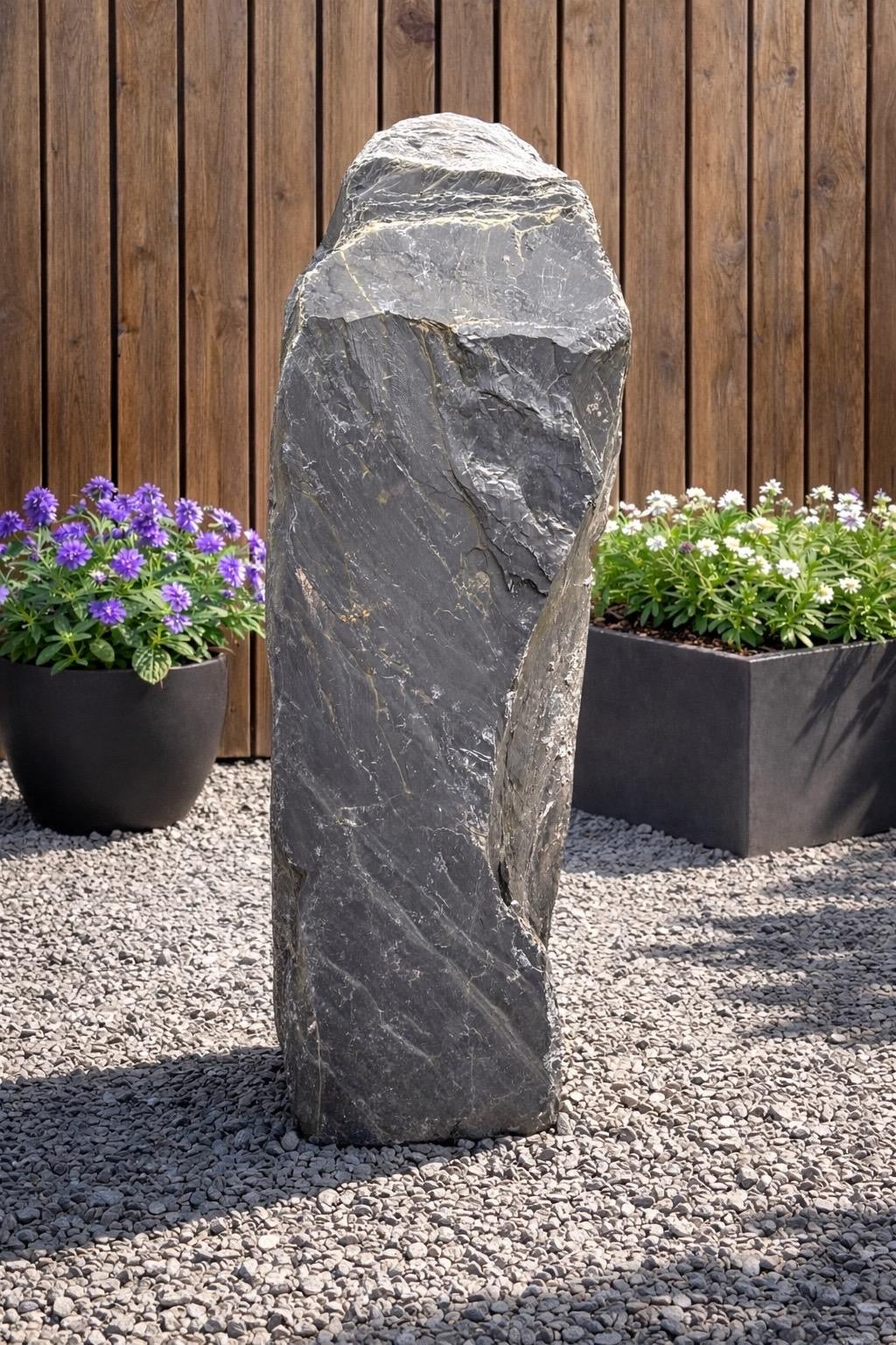 Slate monolith water feature by Water Feature 101, standing among gravel, with purple and white flowers in the background.