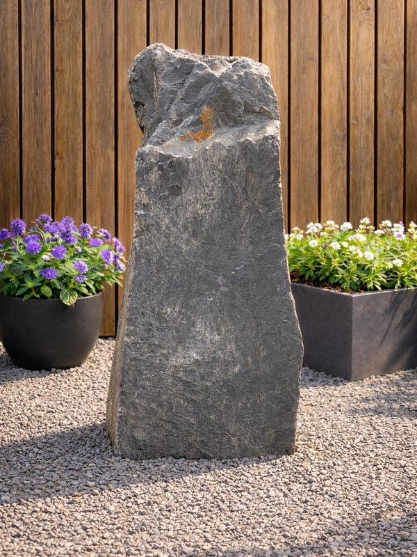 Grey slate monolith water feature from Water Feature 102, standing on a gravel surface, surrounded by potted plants.