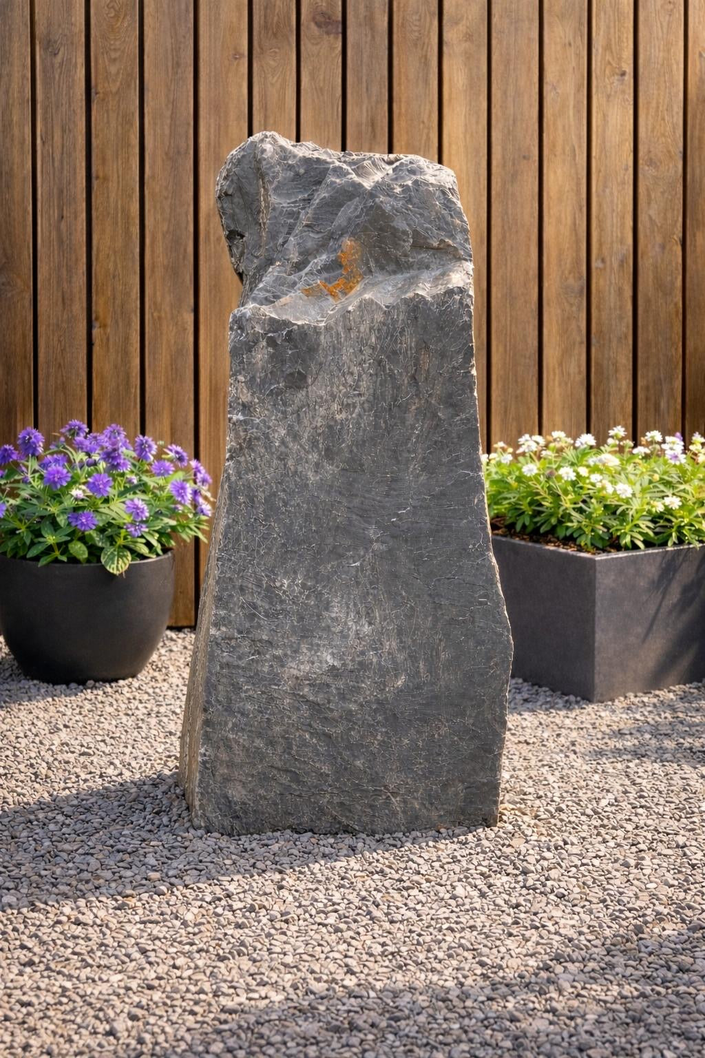 Grey slate monolith water feature from Water Feature 102, standing on a gravel surface, surrounded by potted plants.