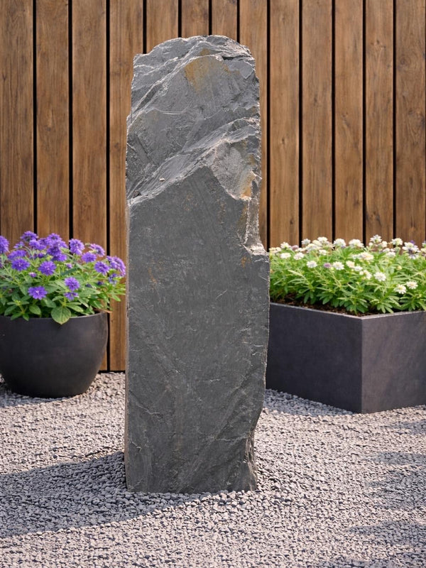 Slate monolith water feature by Water Feature 104, standing tall amongst gravel, next to potted flowers and a wood fence.
