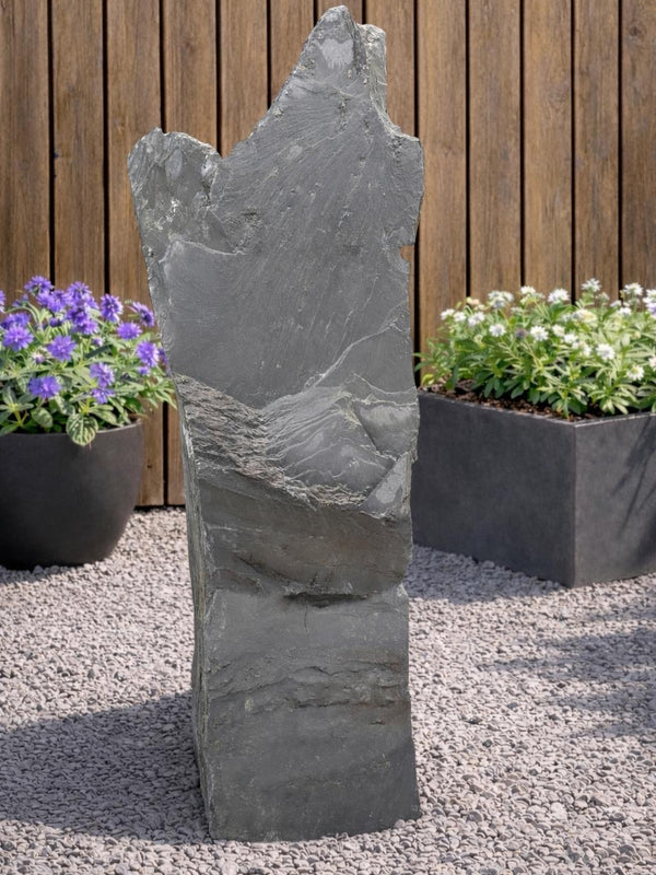 Gray slate monolith by Water Feature 111, standing on gravel between plant containers and a wooden fence.