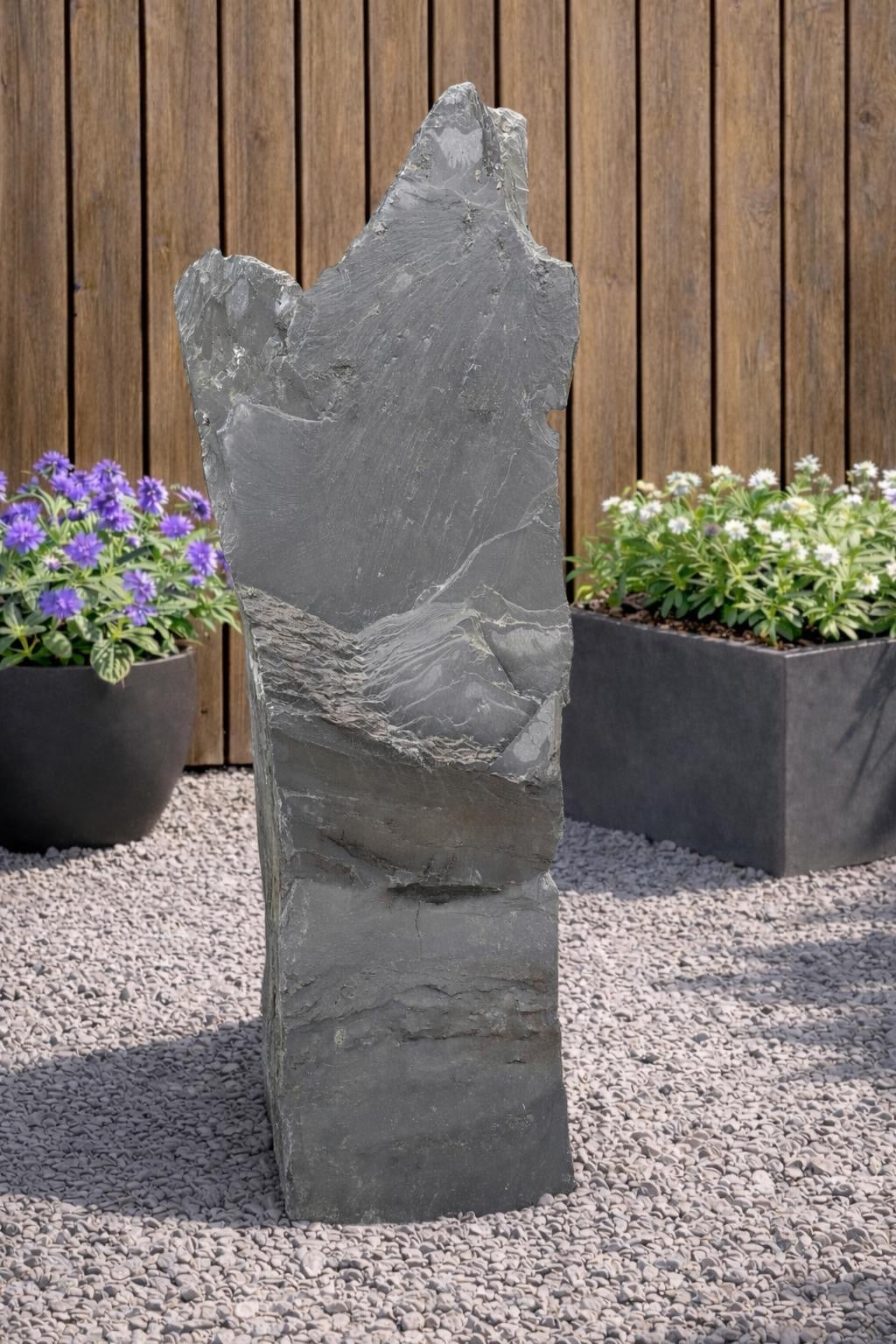 Gray slate monolith by Water Feature 111, standing on gravel between plant containers and a wooden fence.