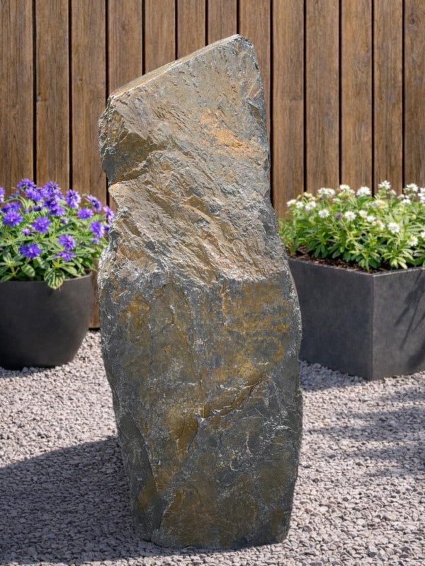 Slate monolith water feature from 108, with a textured surface, placed amongst gravel and plants.