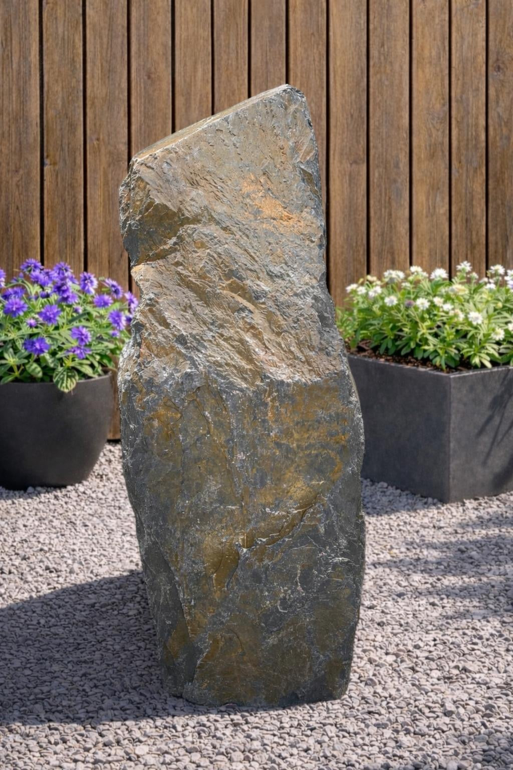 Slate monolith water feature from 108, with a textured surface, placed amongst gravel and plants.