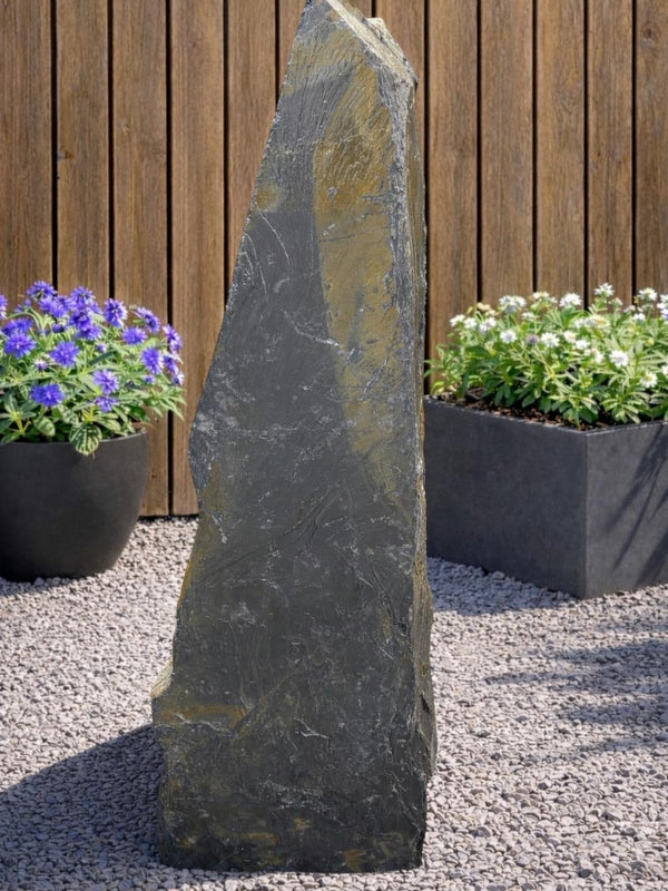 Grey slate monolith standing stone with a pointed top, standing on gravel, with planted containers in the background.