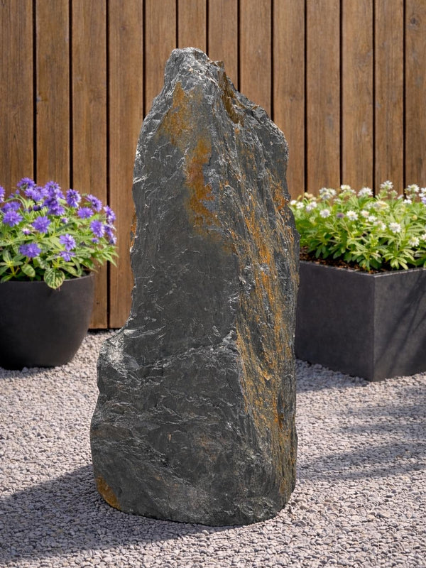 Tall slate monolith water feature with orange streaks, standing on gravel in an outdoor garden setting.