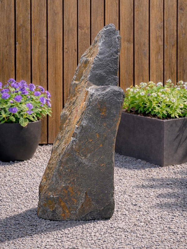 Slate monolith water feature, 1300mm, with hints of orange, set among pebbles with potted plants and wooden fence.