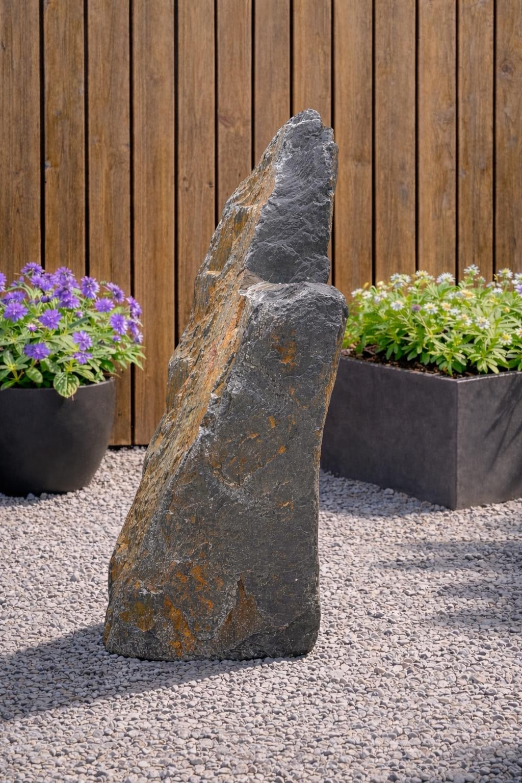 Slate monolith water feature, 1300mm, with hints of orange, set among pebbles with potted plants and wooden fence.