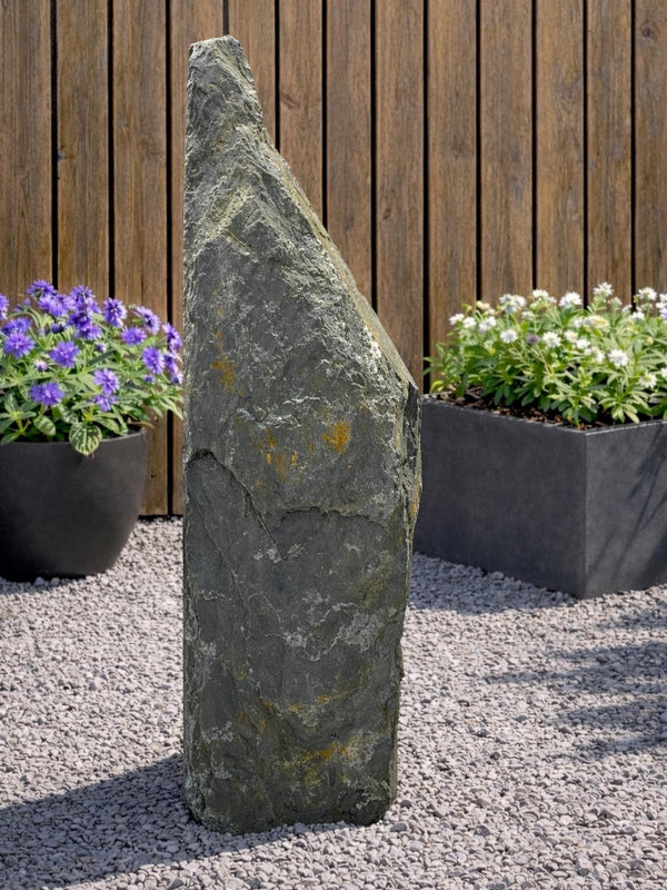 Grey slate monolith water feature by Primrose, standing tall on gravel with garden foliage in the background.
