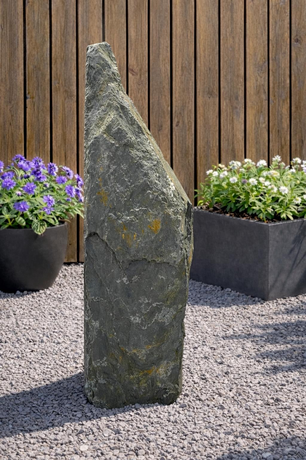 Grey slate monolith water feature by Primrose, standing tall on gravel with garden foliage in the background.
