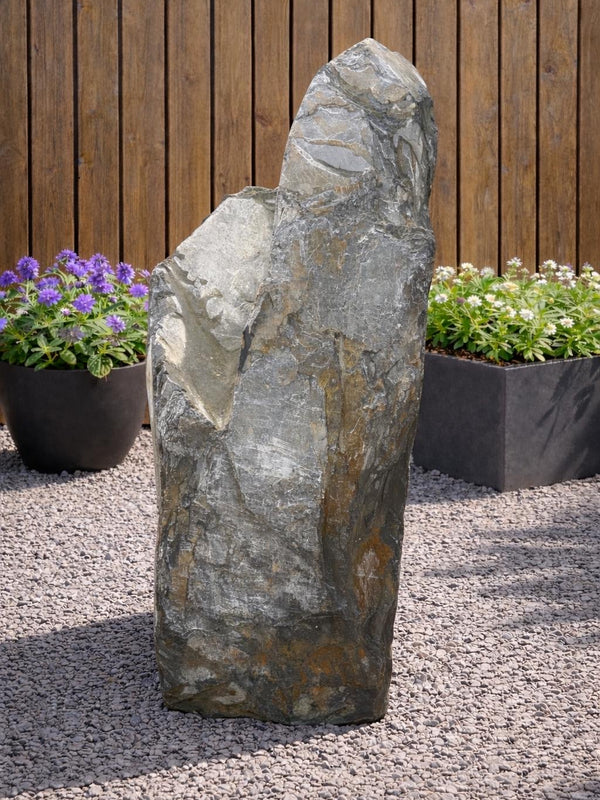 Grey slate monolith water feature from Water Feature 100, set amidst gravel and surrounded by potted flowers.
