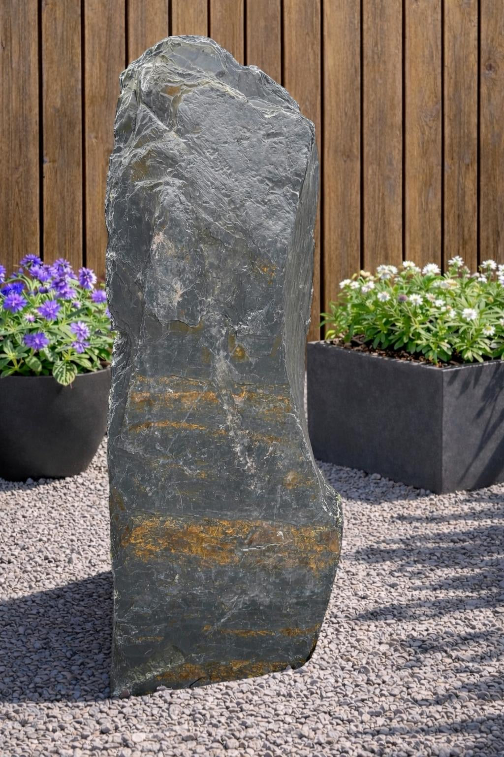 Grey slate monolith standing stone by 132 with gold color striations, set on gravel.