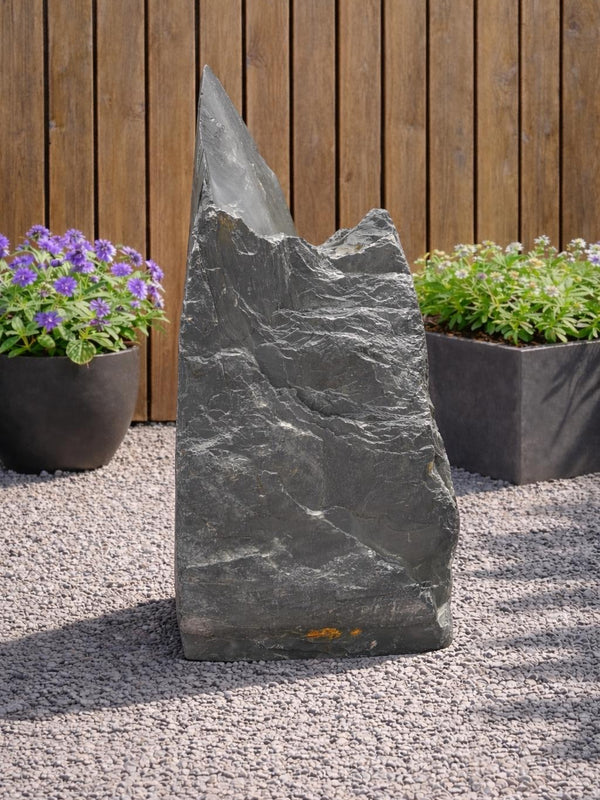 Slate monolith feature stone by 131, standing upright amongst pea gravel, with plants and wood fencing behind.
