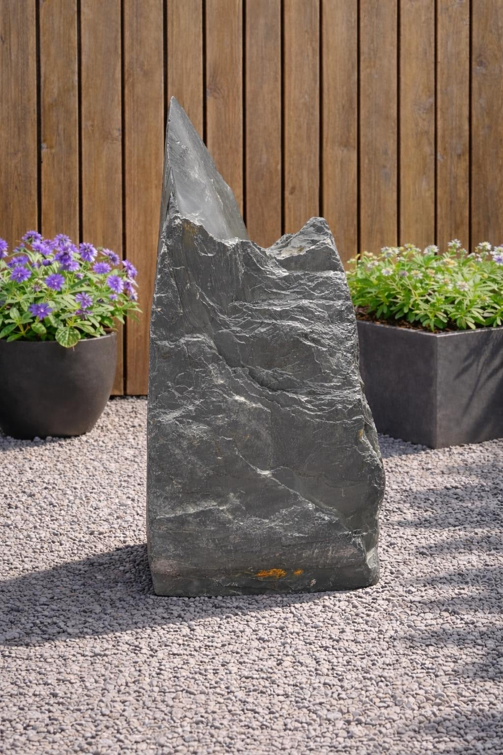 Slate monolith feature stone by 131, standing upright amongst pea gravel, with plants and wood fencing behind.