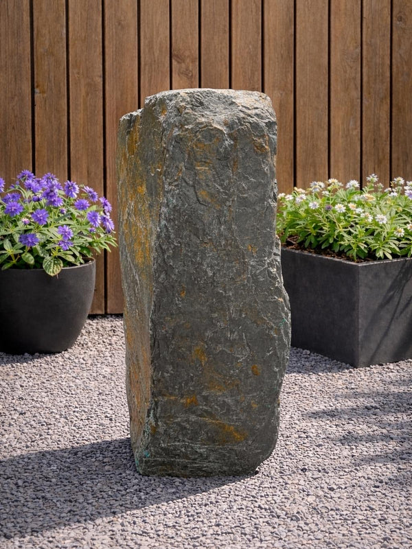 Slate monolith water feature by Water Feature 127, set amongst gravel and surrounded by potted plants.