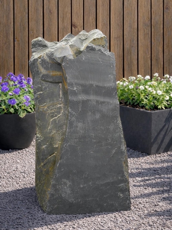 Slate monolith water feature with rough texture, set on pebbles, with planters and wood fence behind.
