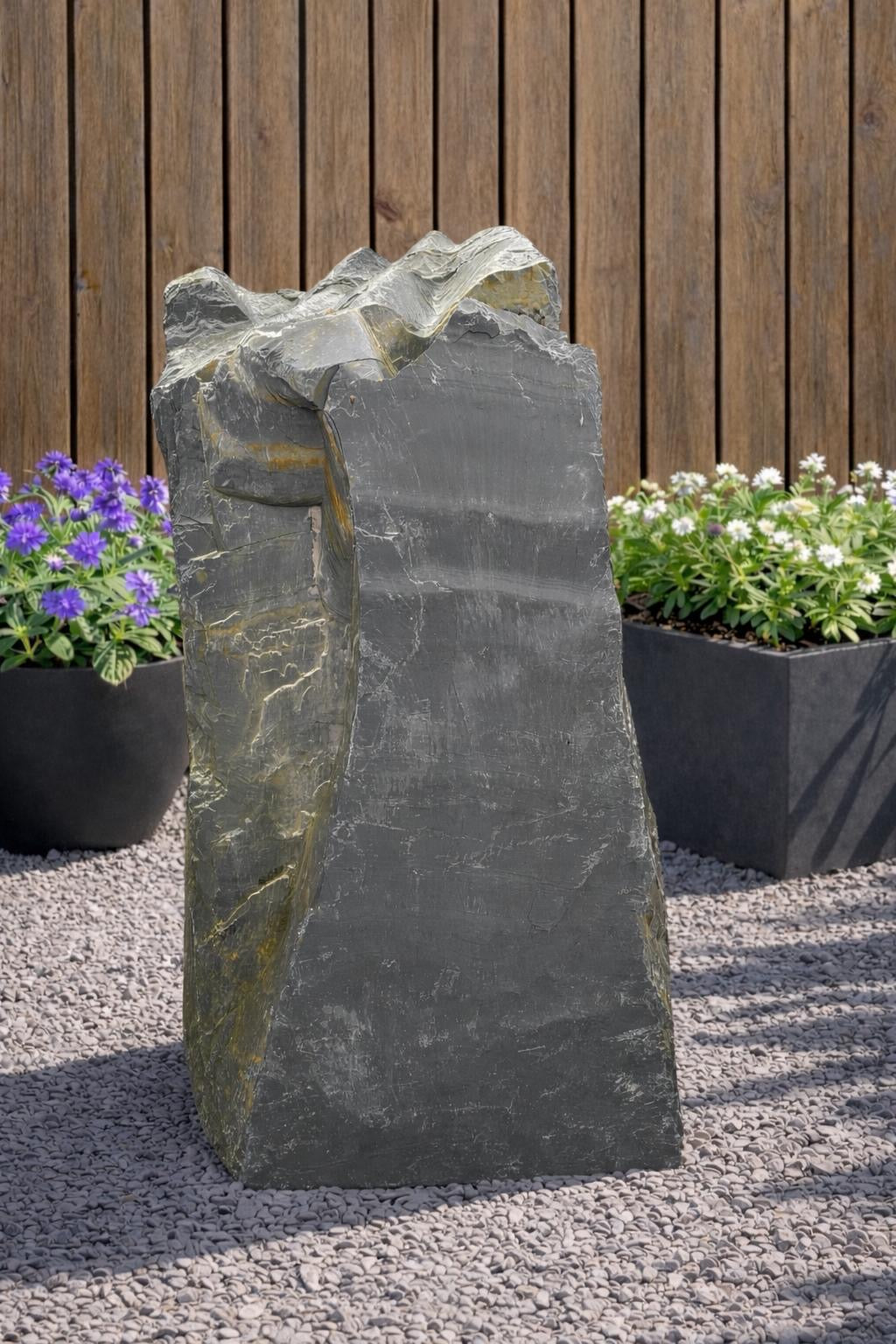 Slate monolith water feature with rough texture, set on pebbles, with planters and wood fence behind.
