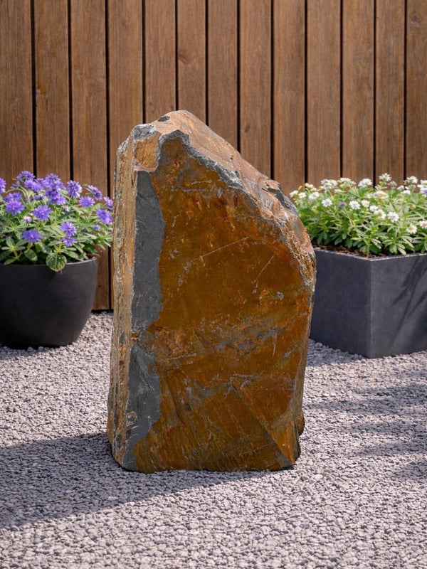 Slate monolith water feature by Water Feature 125, with shades of orange and grey, standing on a gravel surface.
