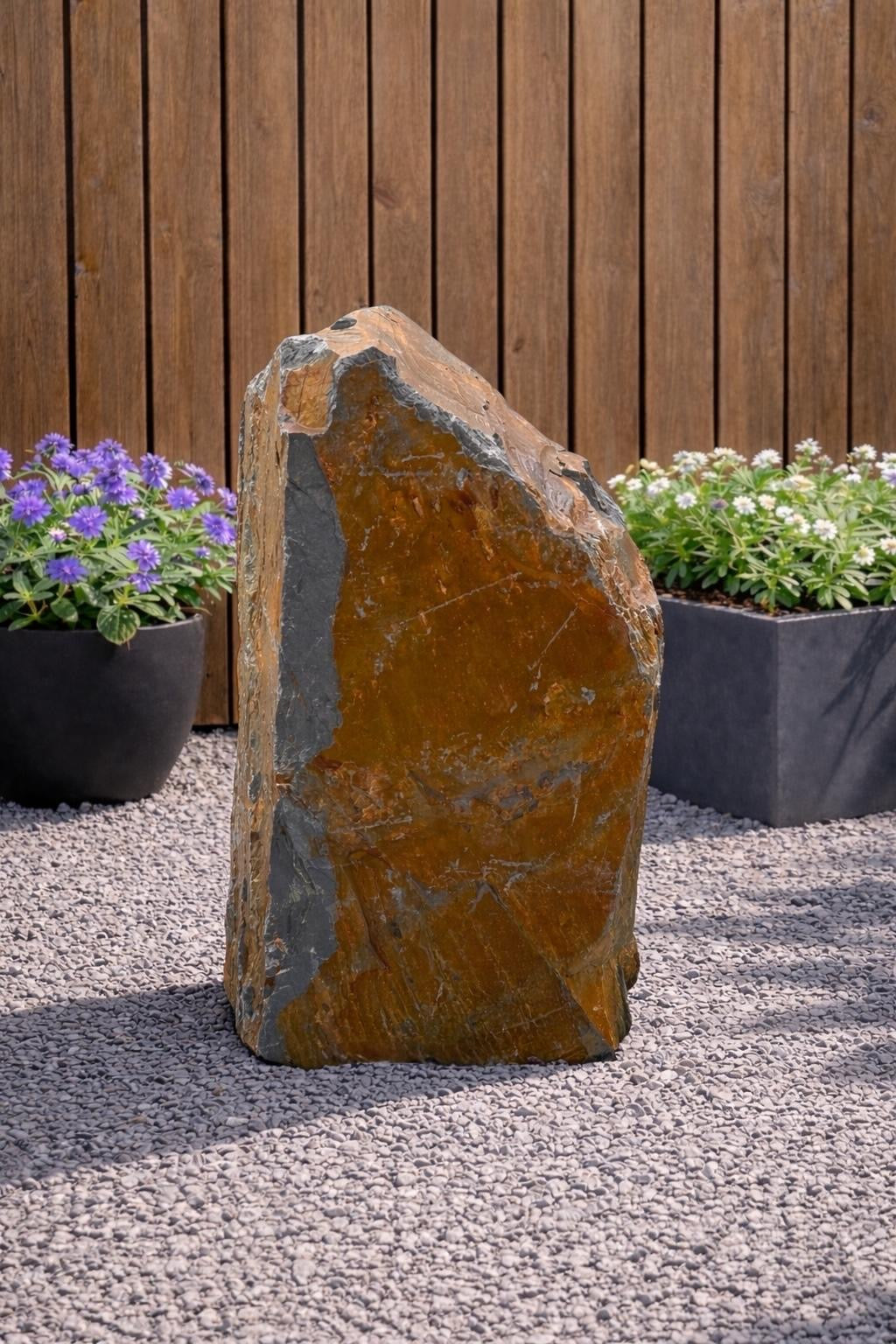 Slate monolith water feature by Water Feature 125, with shades of orange and grey, standing on a gravel surface.
