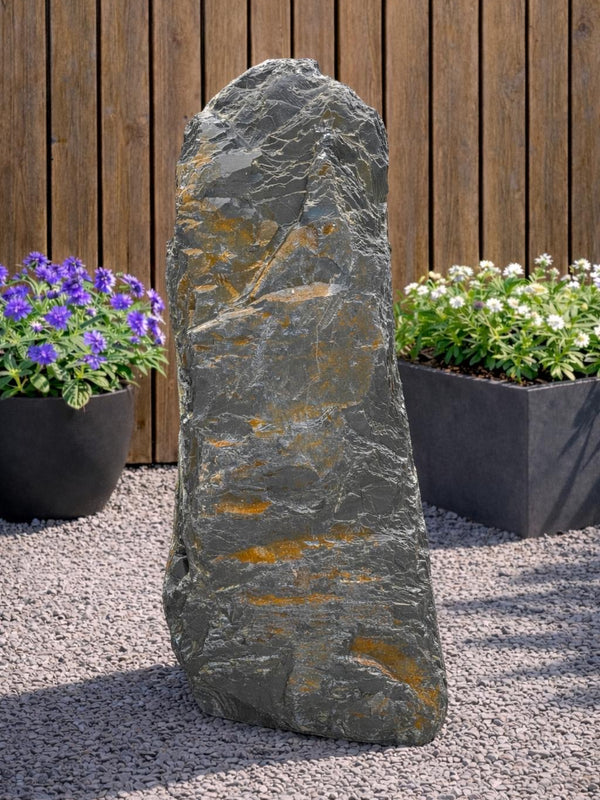 Grey slate monolith standing stone from 123, set amongst gravel with wood paneling and potted flowers in the background.

