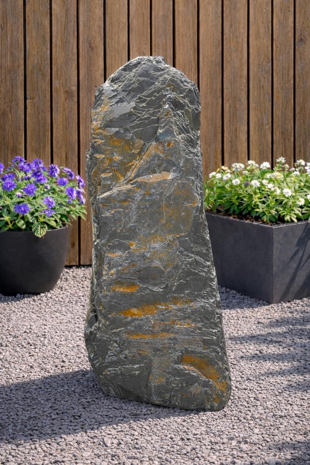 Grey slate monolith standing stone from 123, set amongst gravel with wood paneling and potted flowers in the background.
