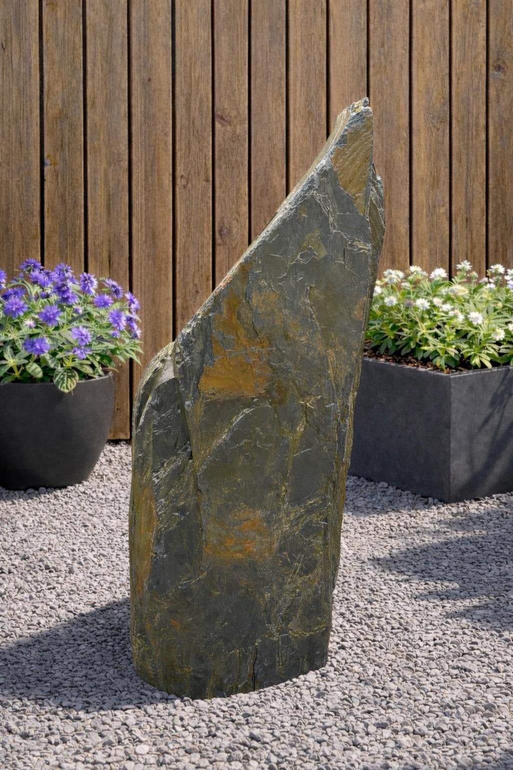 Slate monolith water feature from Water Feature 120 on a gravel surface with purple and white flowers.