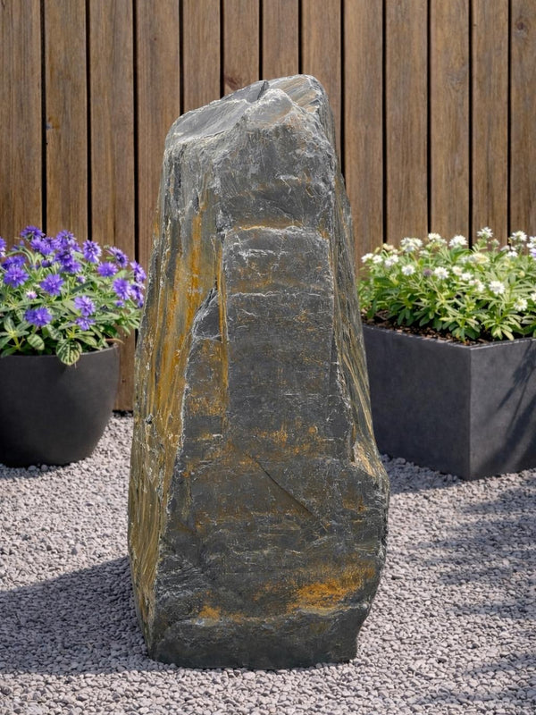Slate monolith water feature with a rough texture, standing on gravel and surrounded by potted plants.