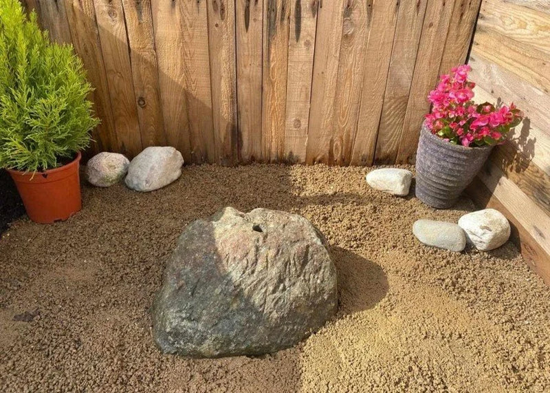 Natural drilled boulder water feature, BWF02, with a hole on top, surrounded by gravel, plants, and stones.