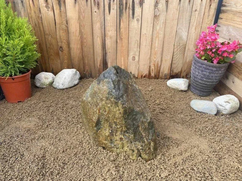 Natural drilled boulder water feature by BWF03, surrounded by pea gravel, plants, and stones.