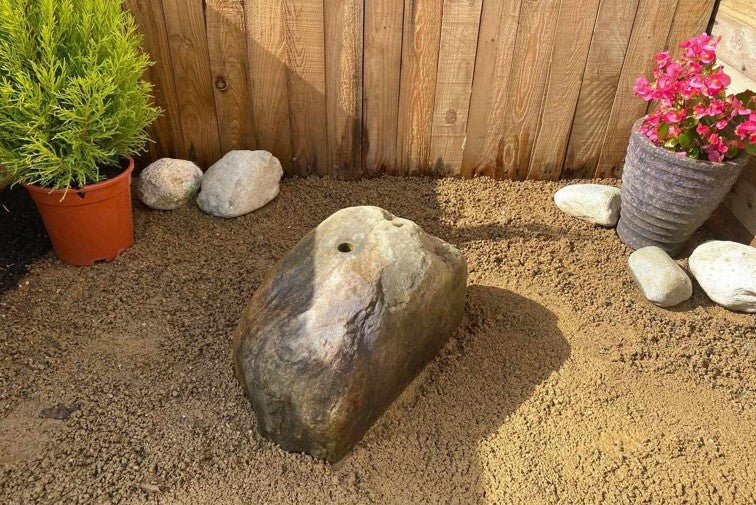 Outdoor scene with a large gray rock featuring a drilled hole, surrounded by gravel, plants, and a wooden fence.