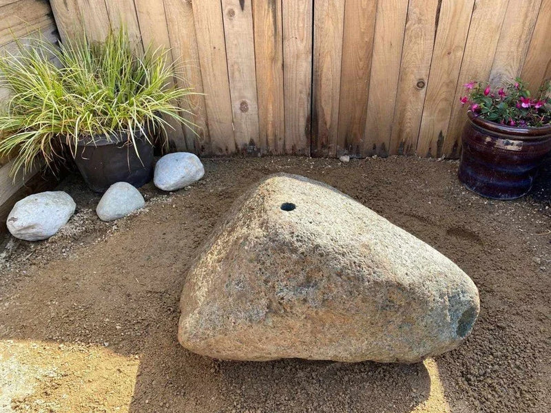 Natural drilled boulder water feature with a centered hole, placed on sandy soil near potted plants and a wooden fence.
