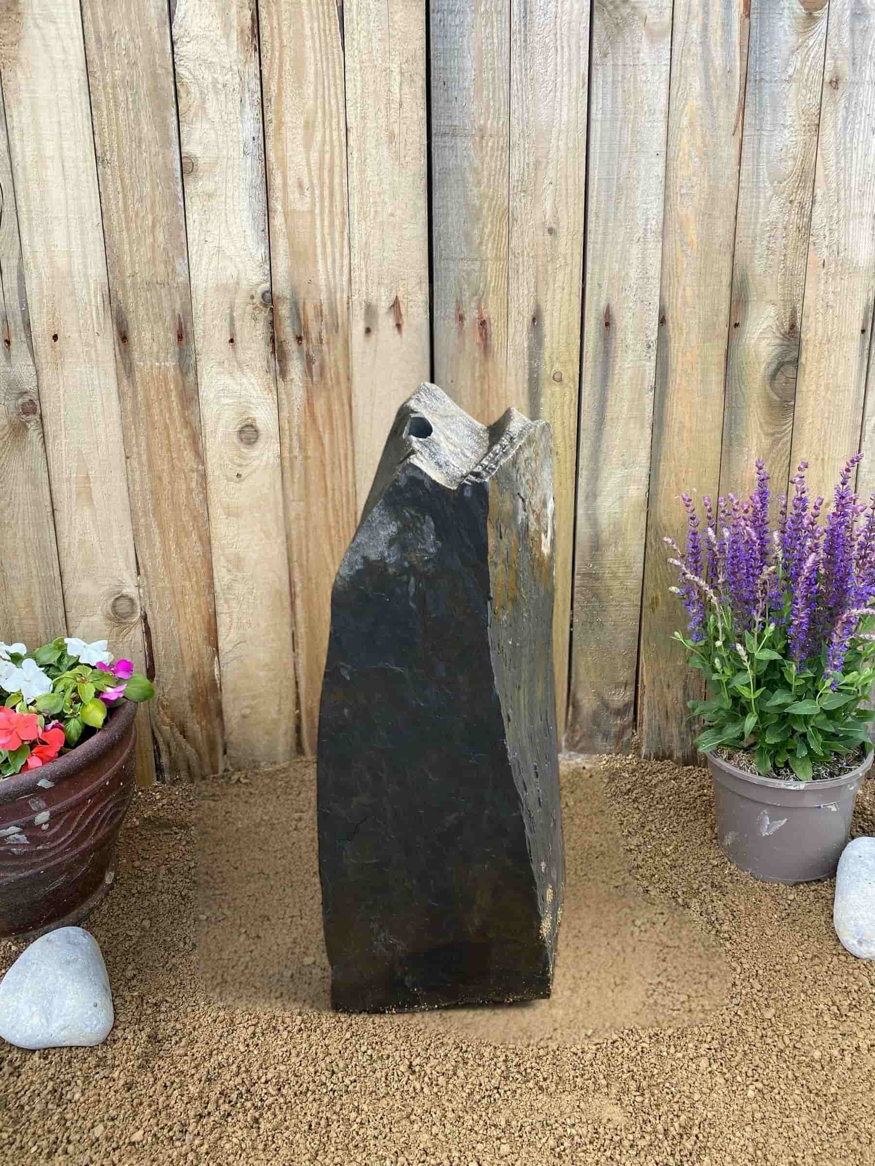 Slate monolith water feature by Water Feature C05 with a drilled hole, standing on gravel near potted flowers.