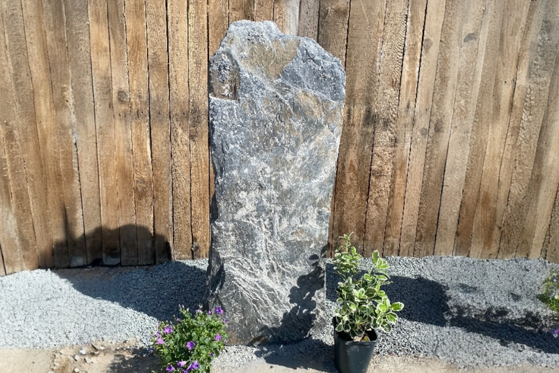 A tall, gray, textured stone slab stands vertically against a wooden fence, flanked by small plants in a sunny outdoor setting.
