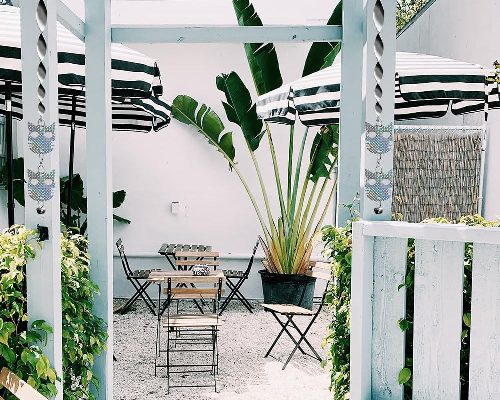 Outdoor patio scene with tables and chairs, striped umbrellas, lush greenery, and whimsical hanging cat decorations. Light and airy atmosphere.
