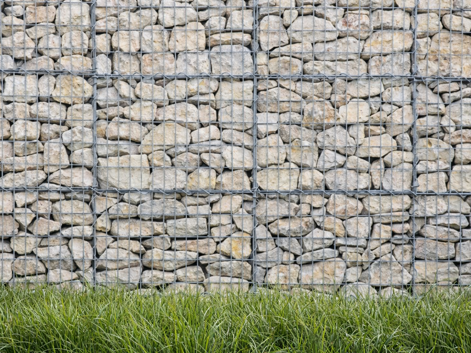 Light limestone gabion stones for gabion baskets, with wire mesh, creating a wall texture, and grass visible along the bottom.
