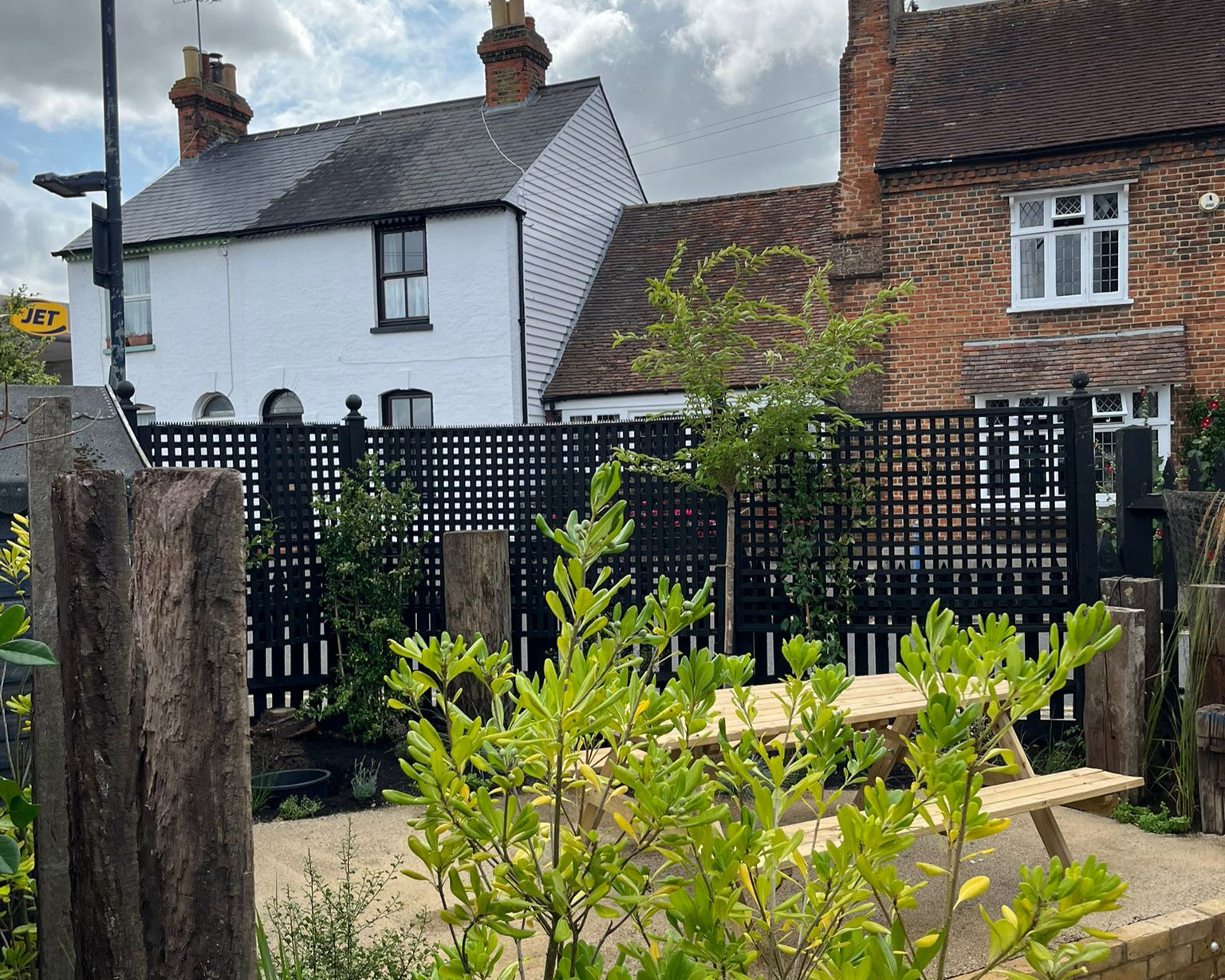Outdoor seating area with a light wood picnic table, framed by greenery and a black lattice fence, set against brick and white buildings under a cloudy sky.
