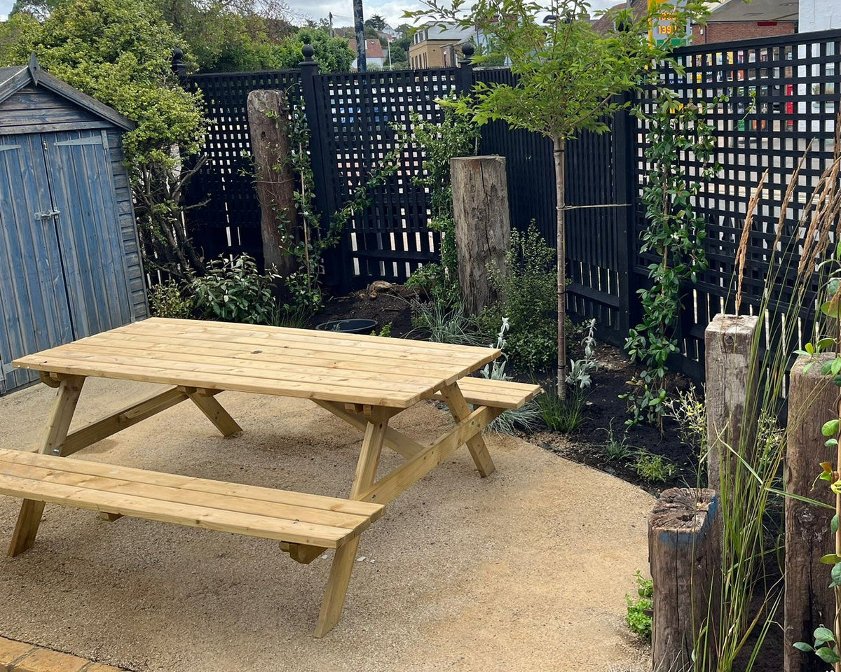 A sunlit backyard scene with a wooden picnic table on gravel, bordered by a black lattice fence, green plants, and wooden posts.
