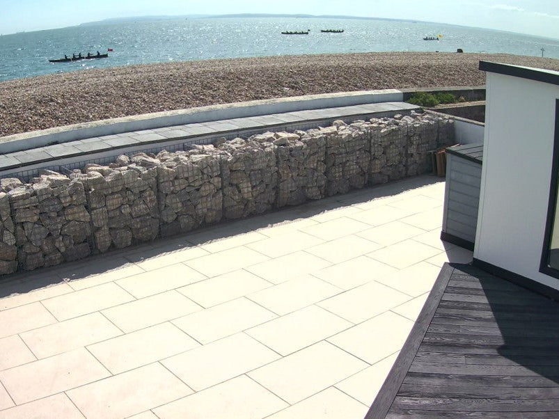 View of a paved patio and rock wall barrier overlooking a stony beach and ocean. Rowers in boats are visible in the distance.
