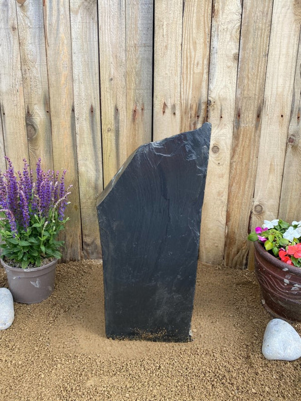 Dark slate monolith water feature by unknown vendor, displayed outdoors with plants and a wooden fence.