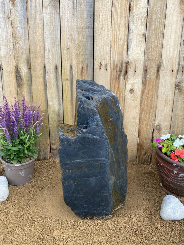 Dark slate monolith water feature by Water Feature C03 standing outdoors between potted flowers and a wooden fence.