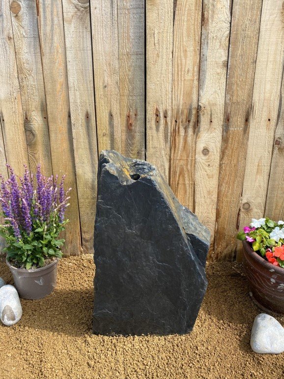 Black slate monolith water feature from C018 with a drilled hole on top, set amongst gravel and planters.